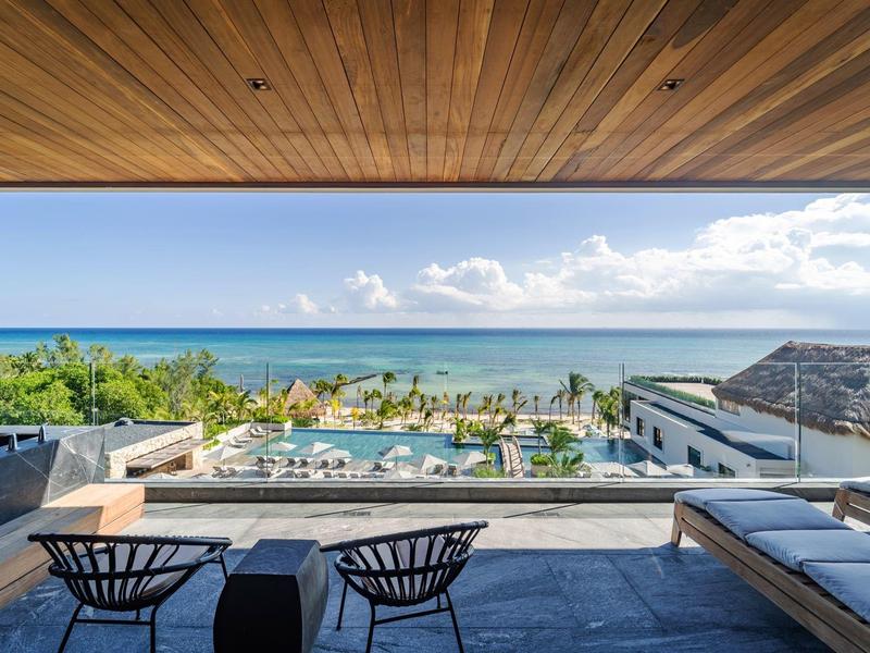 Blick von moderner Terrasse mit Stühlen und Sofa auf Pool, Palmen und Meer bei blauem Himmel.