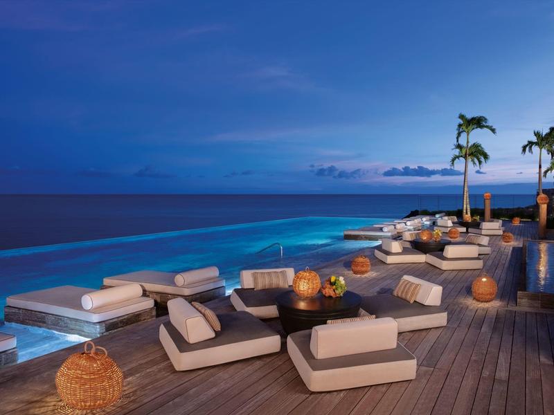 Luxuriöse Terrasse mit modernen Sitzmöbeln, Palmen und stimmungsvoller Beleuchtung am Meer bei Abenddämmerung.