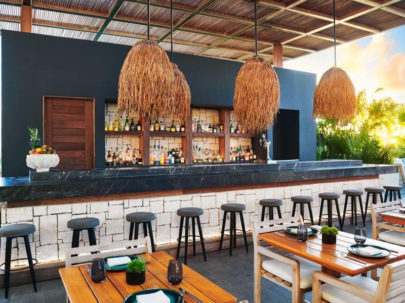 Moderner Außenbarbereich mit Holzmöbeln und dekorativen Lampen unter einem Holzdach bei Sonnenuntergang.