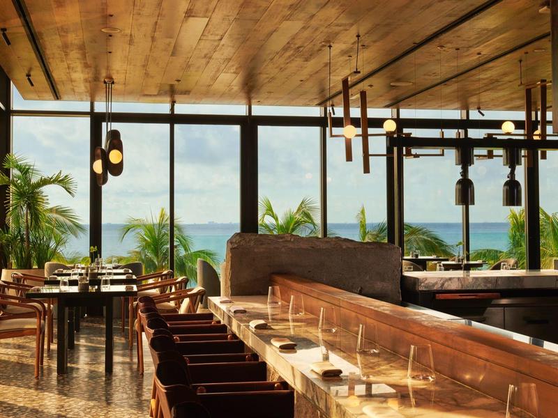 Modernes Restaurant mit Holzdecke und großen Fenstern mit Blick auf Palmen und das Meer.