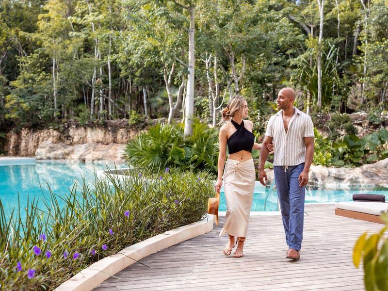 Para spaceruje drewnianym chodnikiem przy wodzie otoczona roślinami.