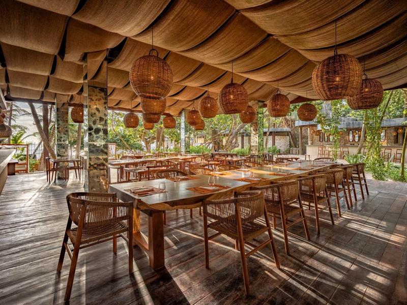 Restauracja na świeżym powietrzu z długimi drewnianymi stołami, krzesłami i dekoracyjnymi lampami pod tkaninowym dachem.