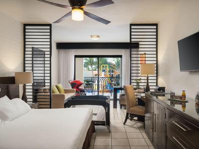 Helles Hotelzimmer mit Doppelbett, Schreibtisch, Fernseher und Balkon mit Blick ins Grüne.