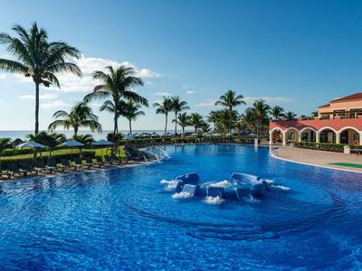 Großer Hotelpool mit Liegestühlen und Palmen am Meer bei klarem Himmel.