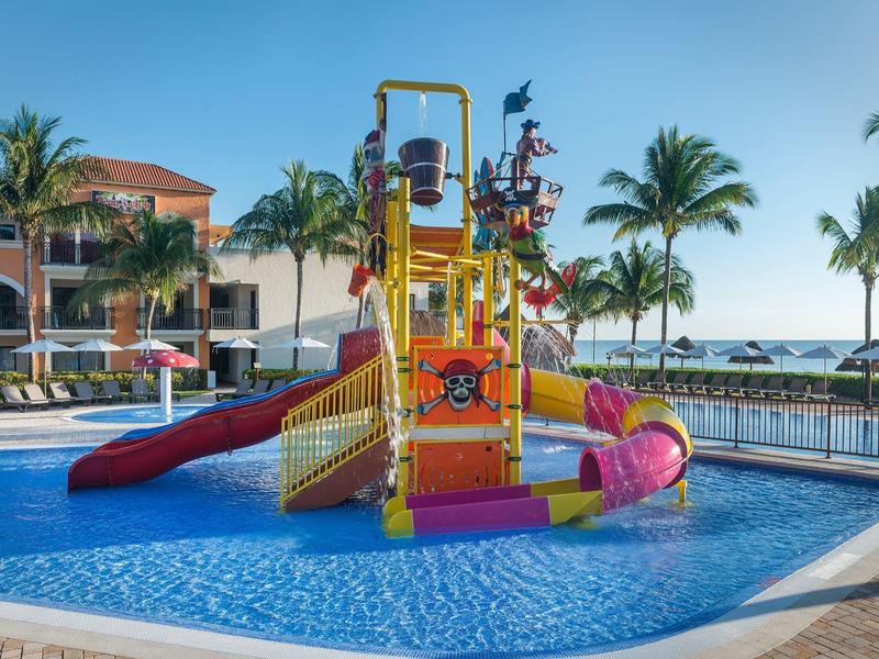 Bunter Wasserspielplatz mit Rutschen in einem flachen Pool, umgeben von Palmen und Hotelgebäuden.