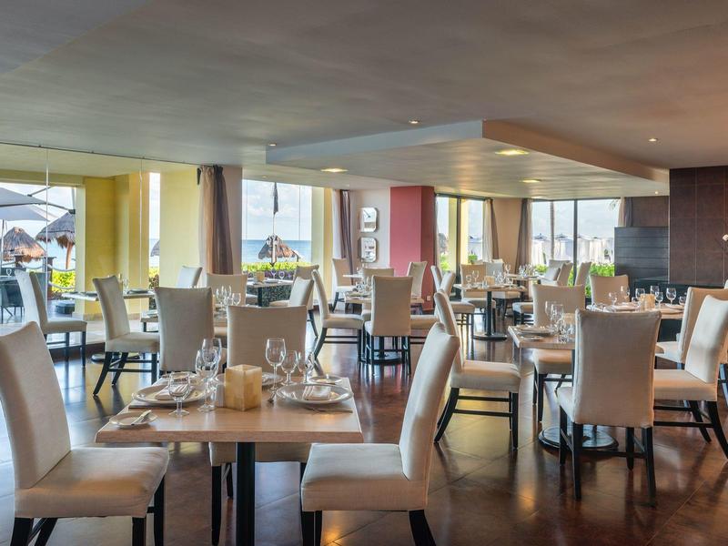 Modernes, helles Restaurant mit weißen Sesseln, gedeckten Tischen und großen Fenstern mit Meerblick.