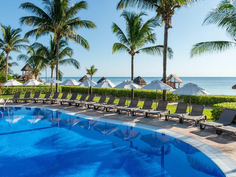 Großer Pool mit blauer Wasserfläche, Liegestühlen und Palmen am Meer unter klarem Himmel.