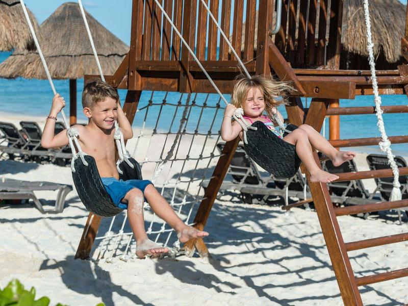 Zwei Kinder schaukeln auf Holzschaukeln am Strand mit weißem Sand und türkisfarbenem Wasser.