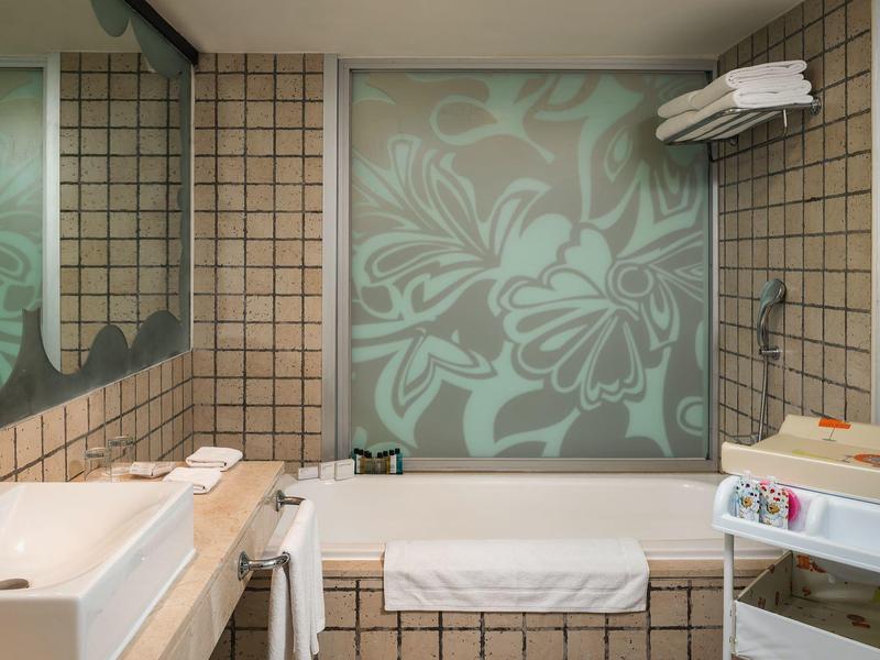 Kleines Badezimmer mit braunen Fliesen, integrierter Badewanne, großer Spiegel und Handtüchern.