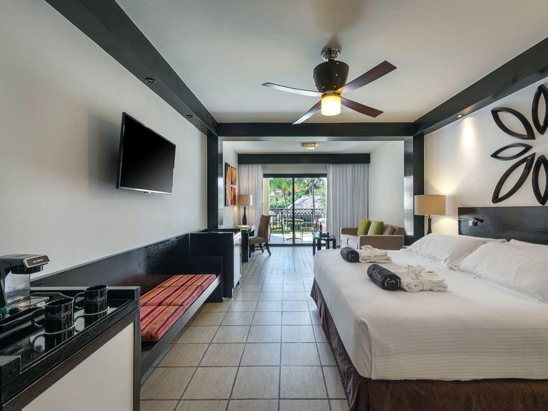 Modernes Hotelzimmer mit Doppelbett, Fernseher, Ventilator und Balkon mit Blick ins Grüne.