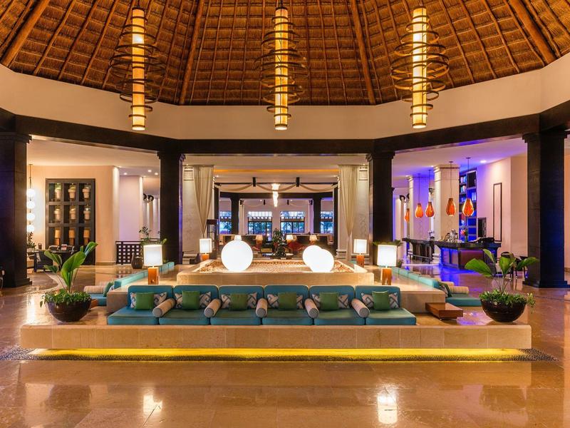 Großer, heller Lobbybereich mit Runddach, Sofas, Pflanzen und moderner Beleuchtung in warmen Tönen.