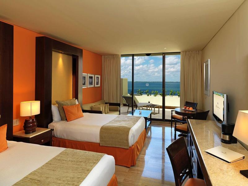 Habitación de hotel luminosa con dos camas, escritorio y vista al mar a través de un gran ventanal.