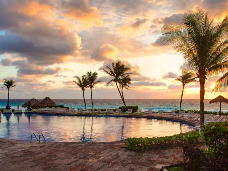 Palmeras y tumbonas rodean una piscina con vistas al mar al atardecer.