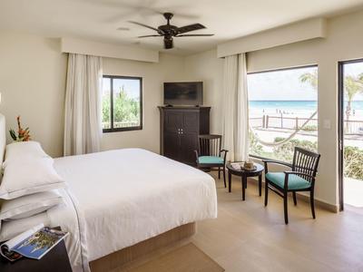 Helles Schlafzimmer mit großem Bett, Ventilator, TV, zwei Stühlen, und Blick auf Strand und Meer.