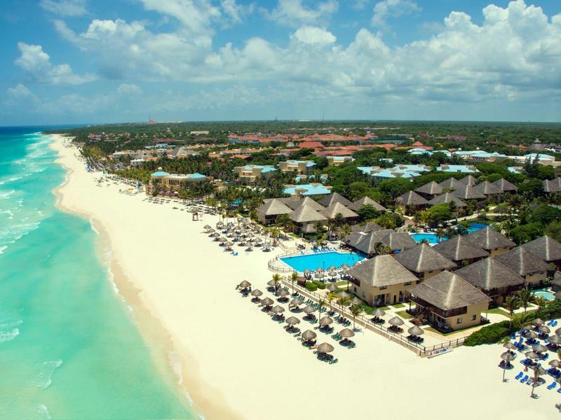 Vue aérienne d'une station tropicale avec plage, piscines et parasols au bord de la mer turquoise.