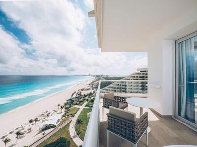 Balcón con silla y mesa ofrece vista al mar de playa de arena blanca y olas azules bajo cielo nublado.