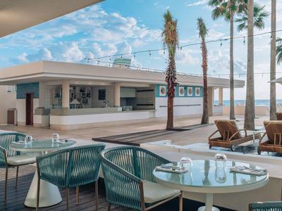 Área exterior moderna con mesas, sillas y barra cerca de la playa bajo cielo azul.