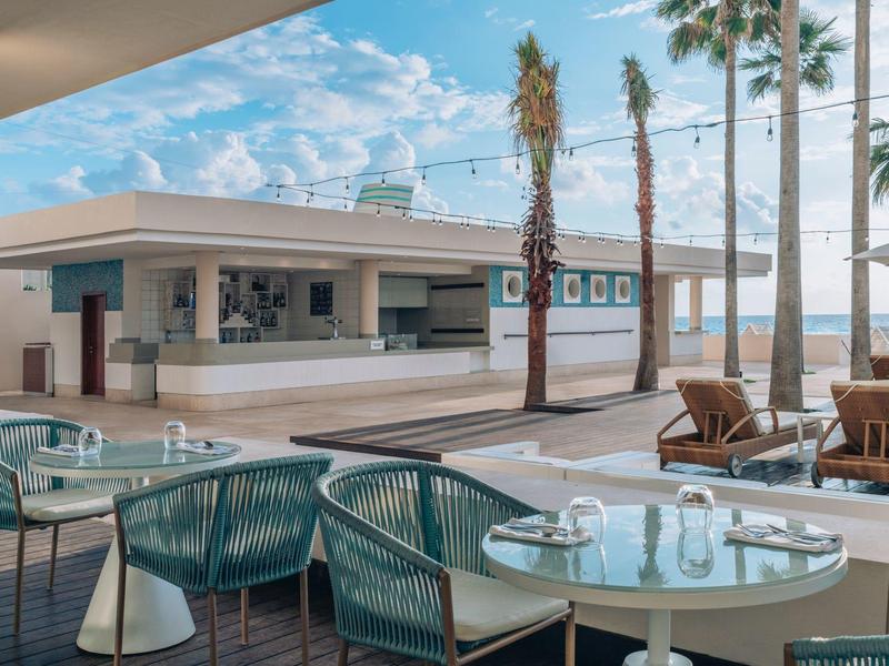 Área exterior moderna con mesas, sillas y barra cerca de la playa bajo cielo azul.