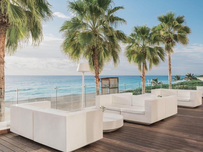Áreas de salón iluminadas con sofás blancos y palmeras en una terraza con vista al mar.