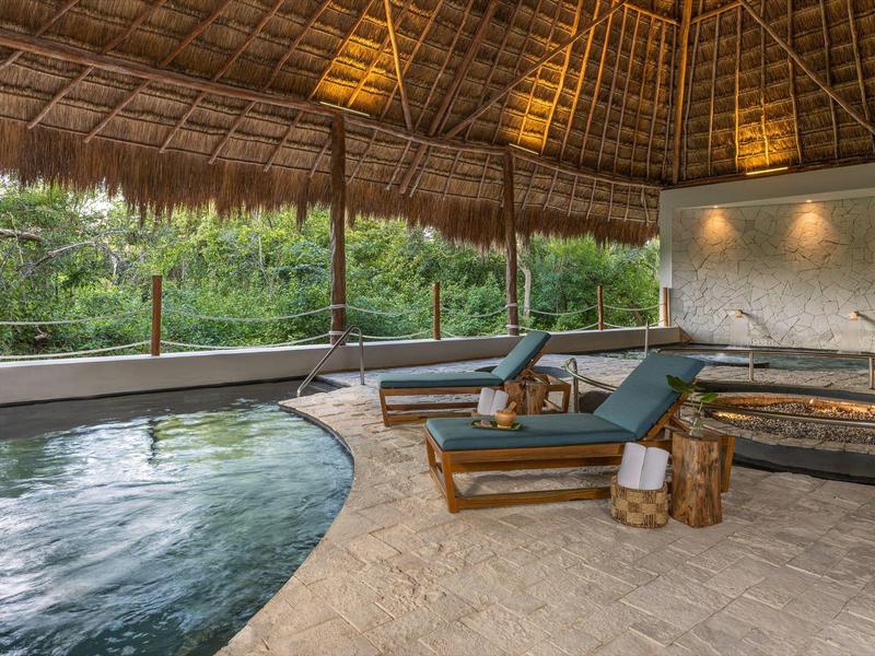 Innenbereich mit runder Schwimmbecken, Holzdach, zwei Liegestühlen und Holzstämmen als Beistelltisch.