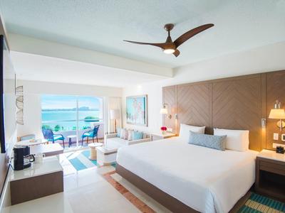 Habitación de hotel luminosa con cama grande, ventilador de techo moderno y vista al mar desde ventanas del piso al techo.