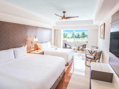 Habitación de hotel luminosa con dos camas grandes y balcón con vistas a palmeras y mar.