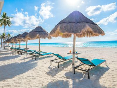 Sandstrand mit türkisblauem Meer, blauen Liegestühlen und Strohhut-Sonnenschirmen bei Sonnenschein.
