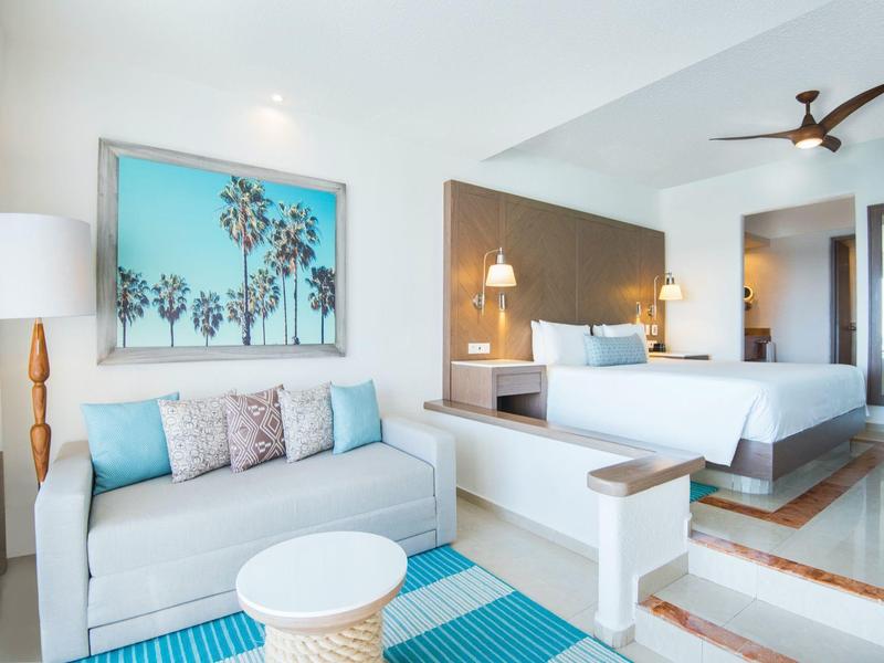 Habitación de hotel luminosa con sofá blanco, alfombra azul y cama grande con ventilador de techo.