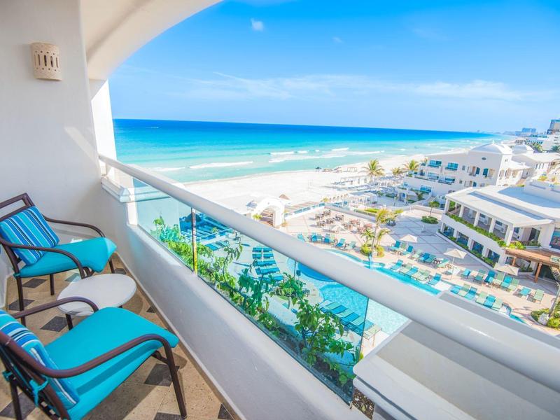 Balcón con sillas azules y vista a la playa, el mar y las instalaciones del hotel bajo un cielo azul.