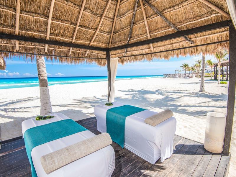 Dos camillas de masaje bajo un techo de paja en la playa de arena blanca con vista al mar azul.