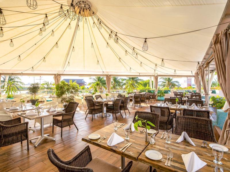 Terraza luminosa del hotel con suelo de madera, mesas puestas y vista a palmeras y mar.