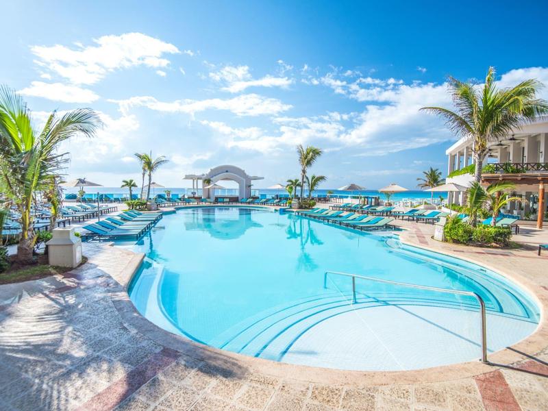 Piscina exterior luminosa con palmeras y tumbonas bajo un cielo azul en un complejo hotelero.