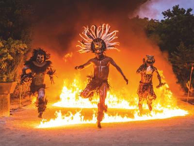 Drei Tänzer in traditioneller Kleidung springen vor einem großen Lagerfeuer unter dunklem Himmel.