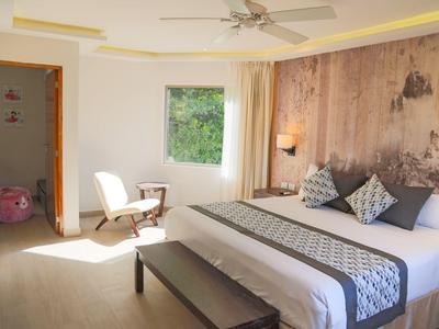 Habitación de hotel luminosa con cama grande, banco al pie, sillas junto a la ventana y pared de madera.