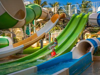 Kind spielt auf grüner Wasserrutsche in tropischem Wasserpark mit mehreren Rutschen.