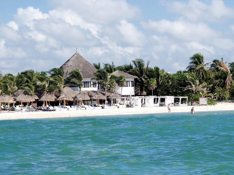 Resort sulla spiaggia con palme, tetti di paglia e acqua turchese sotto un cielo nuvoloso.