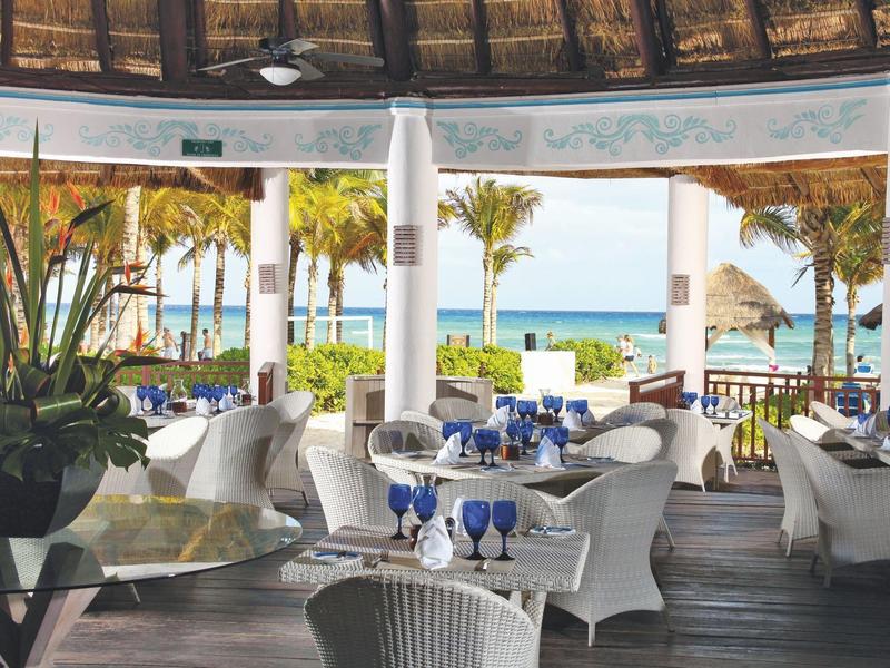 Ristorante elegante con mobili bianchi e vista mare sotto un tetto di paglia.