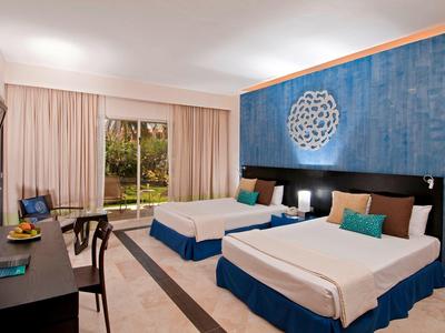 Hotelzimmer mit zwei Doppelbetten, blauem Kopfteil, Schreibtisch und Gartenblick durch Fenster.