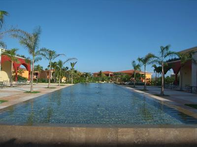 Breiter Pool mit klarem Wasser, Palmen und bunten Häusern rechts und links bei blauem Himmel.
