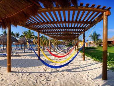 Mehrfarbige Hängematten hängen auf Sand unter einem Holzpergola, umgeben von Palmen und Rasen.