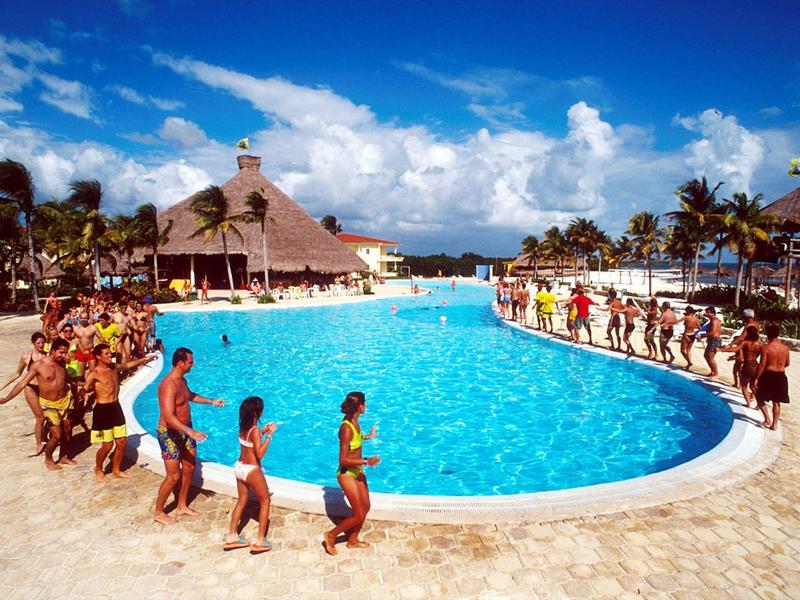 Persone che si godono una grande piscina in un resort tropicale con palme e costruzioni in paglia.