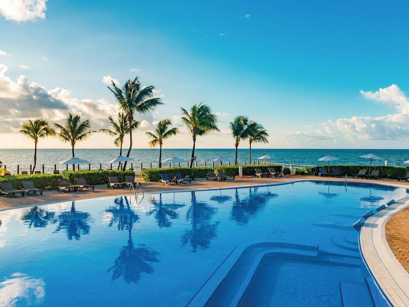 Piscina all'aperto con lettini e palme sul mare sotto un cielo azzurro.