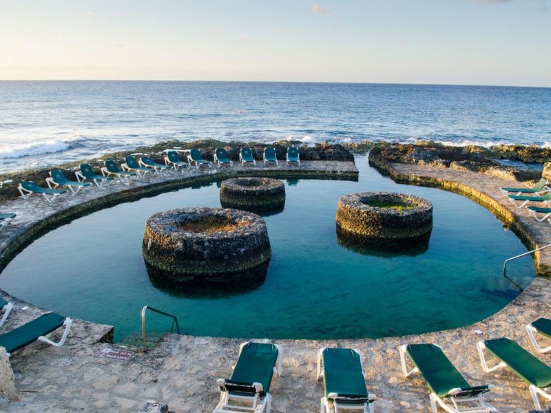 Piscina rotonda di acqua di mare con tre isole circolari e lettini lungo il bordo su mare calmo.