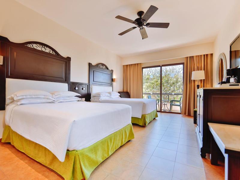 Camera d'hotel luminosa con due letti matrimoniali, ventilatore a soffitto e balcone con vista.