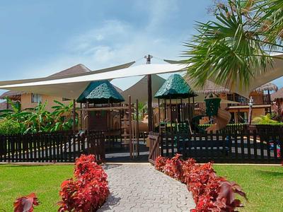 Parco giochi per bambini con attrezzature e tenda da sole, circondato da cespugli e case.