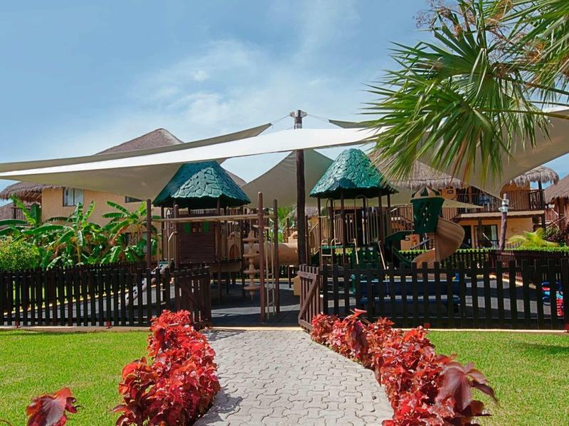 Parco giochi per bambini con attrezzature e tenda da sole, circondato da cespugli e case.