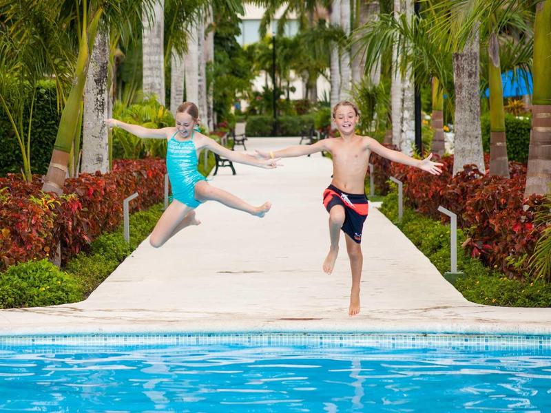 Due bambini saltano con gioia da un sentiero in una piscina in un resort tropicale.