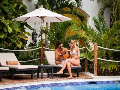 Zwei Frauen sitzen entspannt mit Getränken auf Poolliegen neben einem Pool, umgeben von grünen Pflanzen.