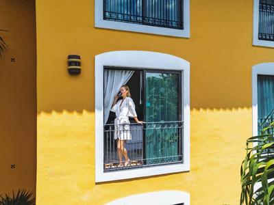 Frau in weißem Kleid auf Balkon eines gelben Hauses mit weißen Fensterrahmen und grünen Geländern.