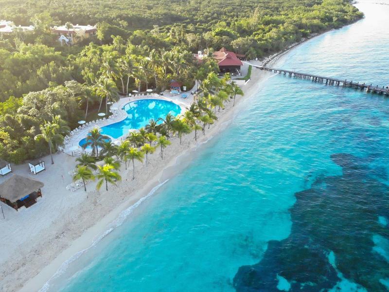 Resort sulla spiaggia con piscina, palme e molo in mare turchese circondato da una fitta vegetazione.